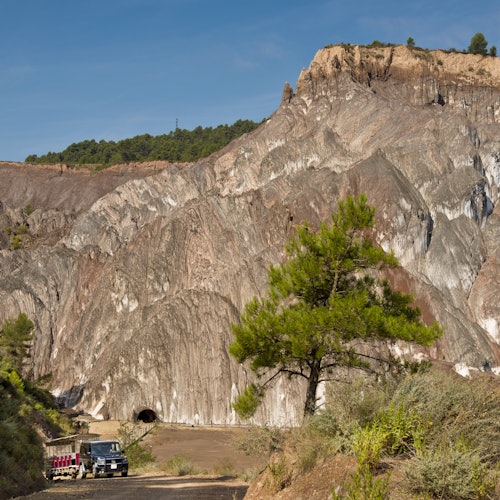 A rugged landscape featuring a large rock mountain, tunnel entrance, green trees, and a parked vehicle under a clear blue sky.
