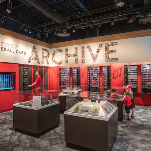 People viewing baseball card displays in a brightly lit archive. Glass cases and wall cabinets showcase various card collections.