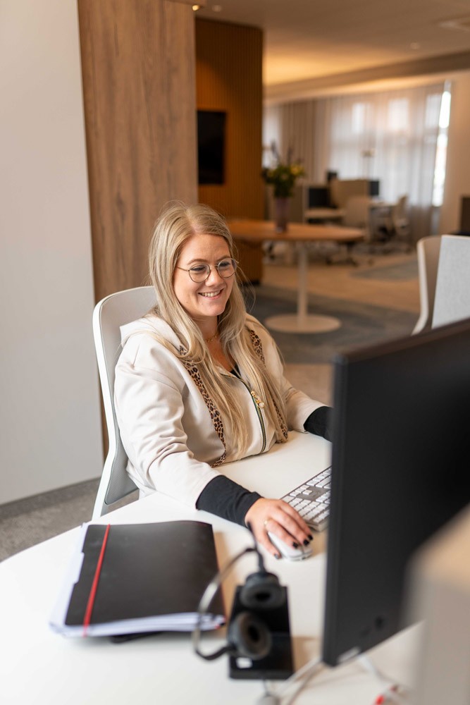 Mitarbeiterin im hellen Blazer sitzt lächelnd an einem Schreibtisch im Büro von SLB.