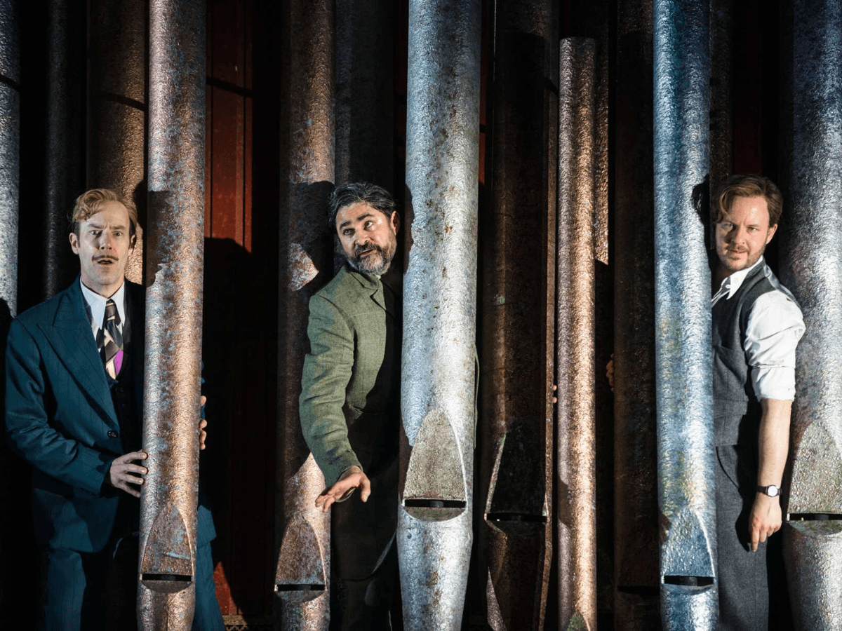 Three men peering between tall metal pipes in a comic moment from Twelfth Night.