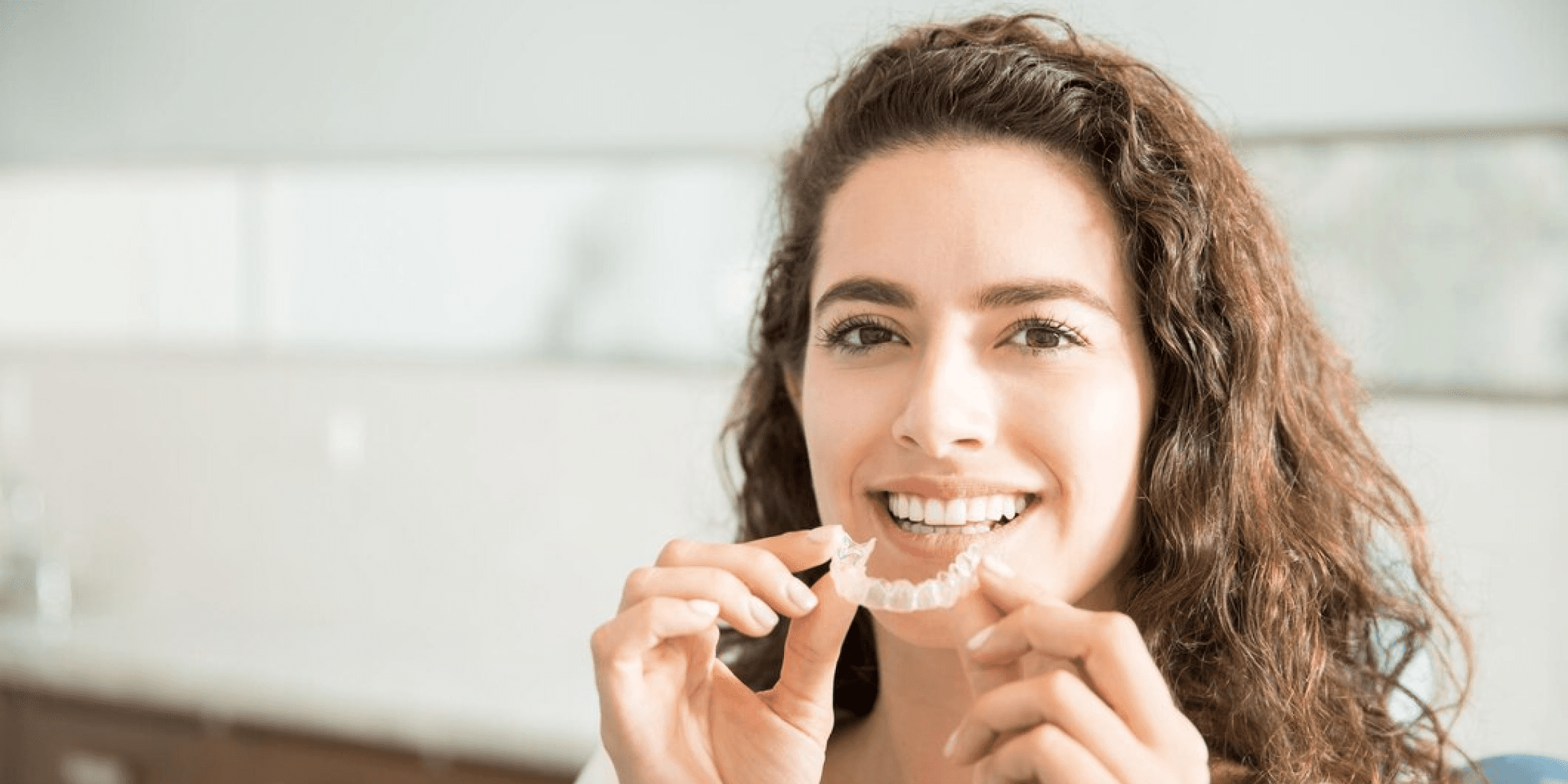 A smiling woman holding a clear dental aligner, illustrating the natural look of inlays that blend seamlessly with existing teeth for a flawless smile.