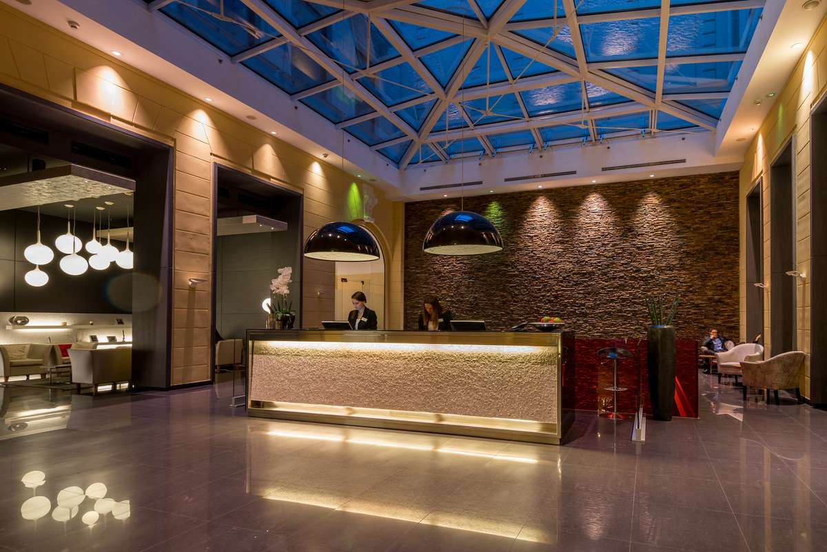 A modern hotel lobby features a long illuminated reception desk under a blue glass ceiling with warm spotlights and stone walls.
