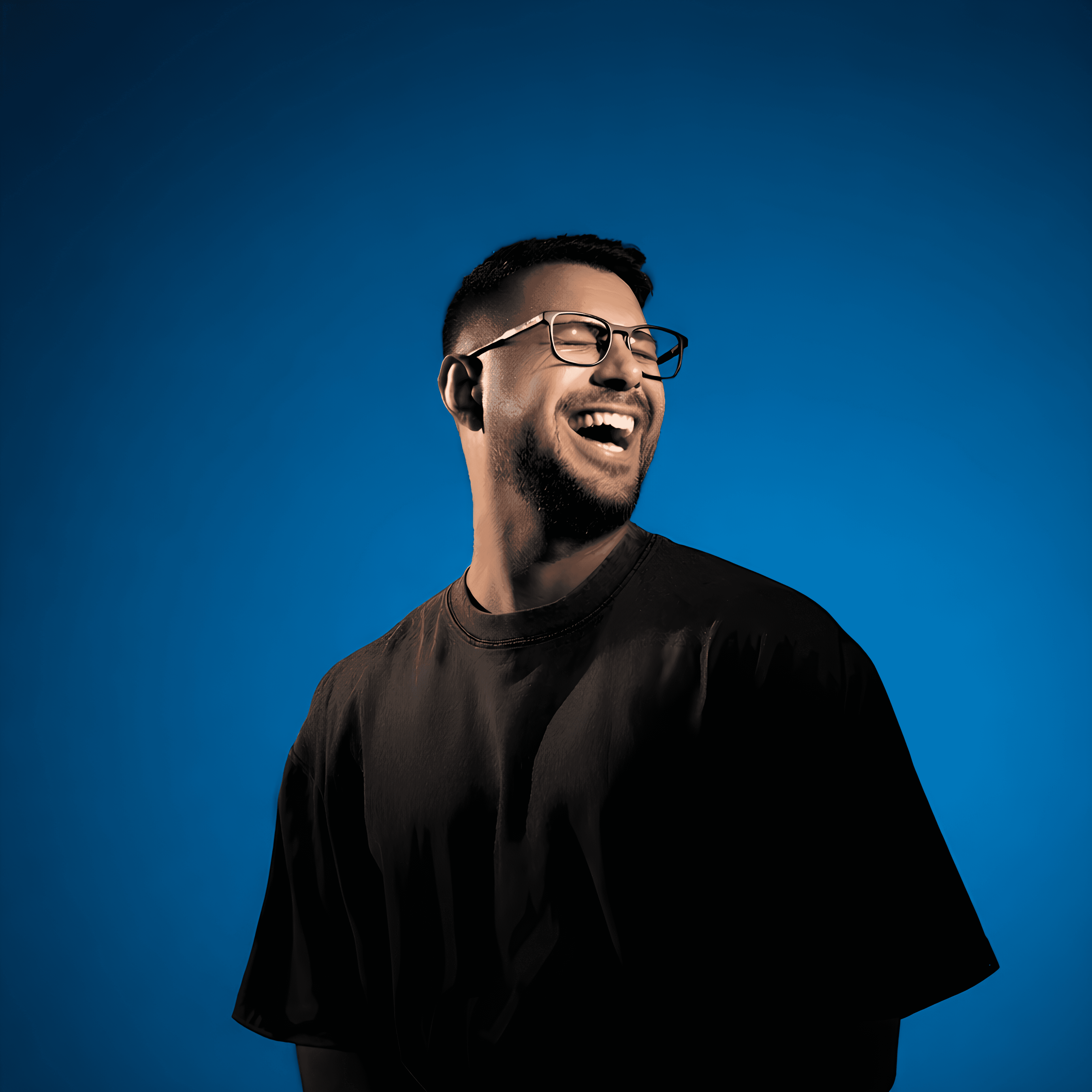 Joyful portrait of a young man with short dark hair and a neatly trimmed beard, laughing heartily with eyes closed behind clear-framed glasses; warm dramatic lighting casts a rich teal-blue glow across his face and oversized dark t-shirt, creating a vibrant, energetic mood against a solid deep blue background.