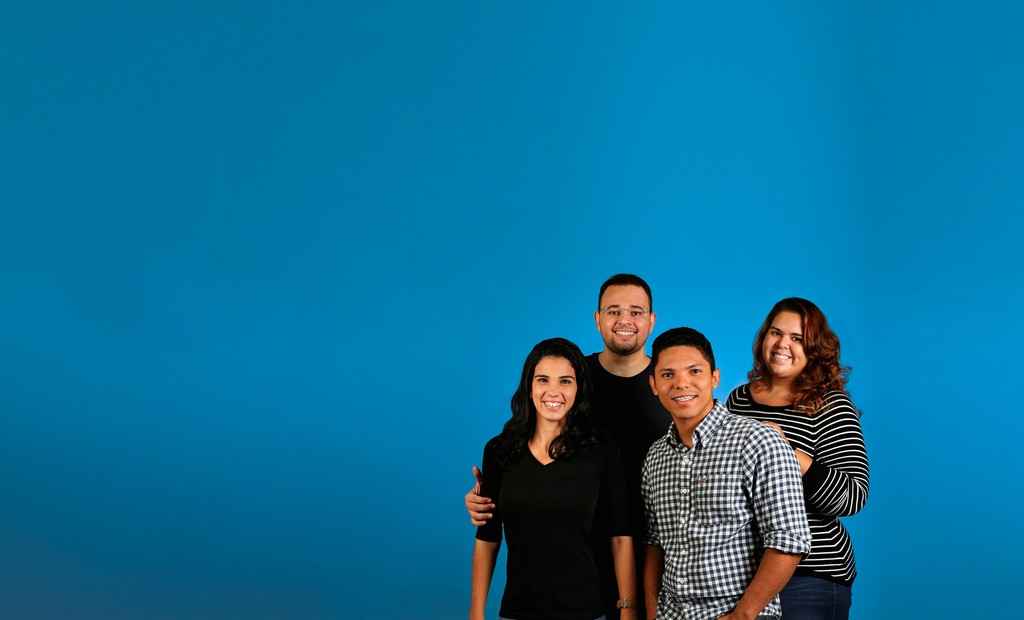 A group of four smiling individuals stands together against a solid blue background. Their casual attire and relaxed poses suggest a friendly and welcoming atmosphere, emphasizing diversity, teamwork, and positive social interaction.