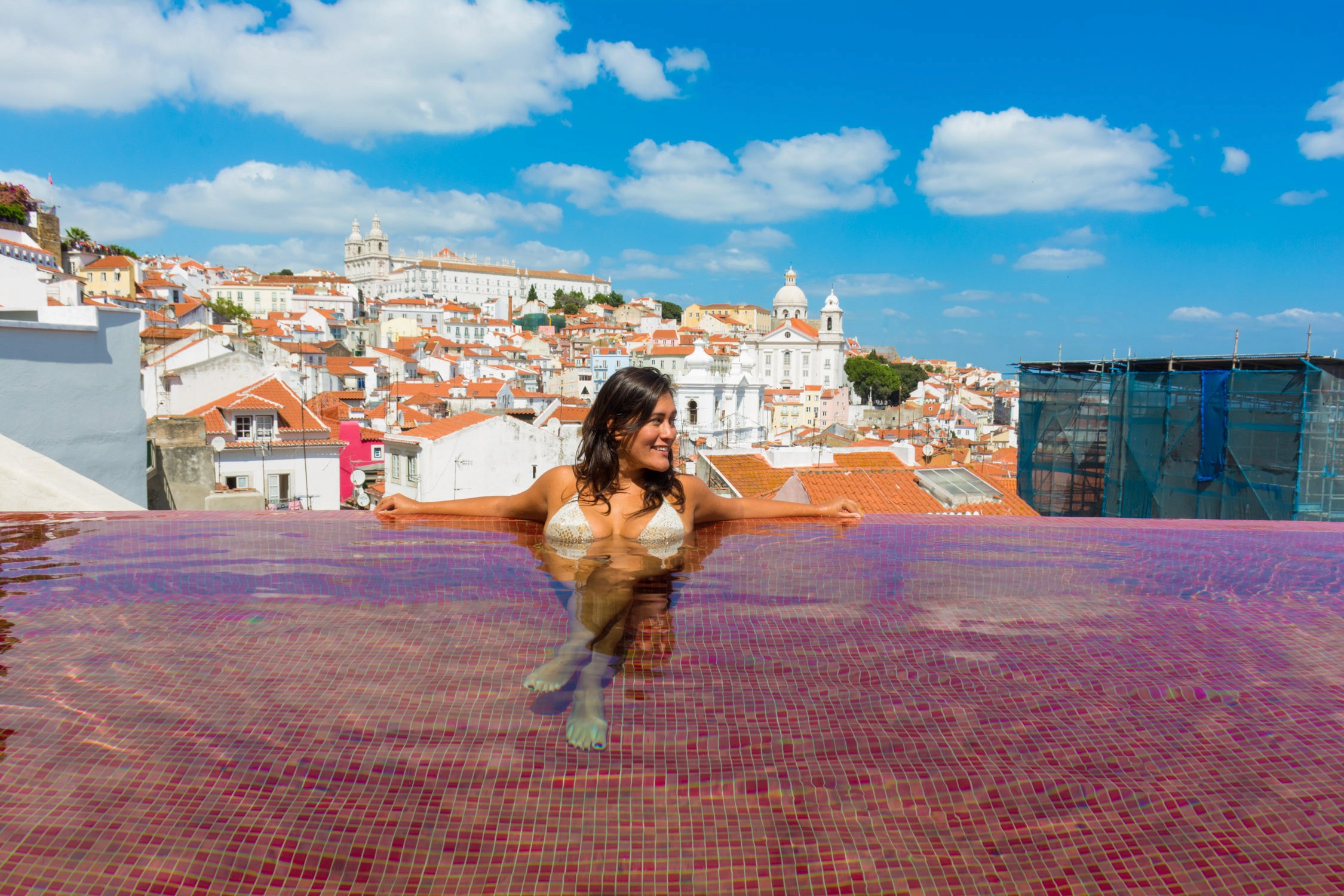 A woman in a bikini poses on a pink tiled terrace overlooking a coastal town with white buildings and blue roofs.