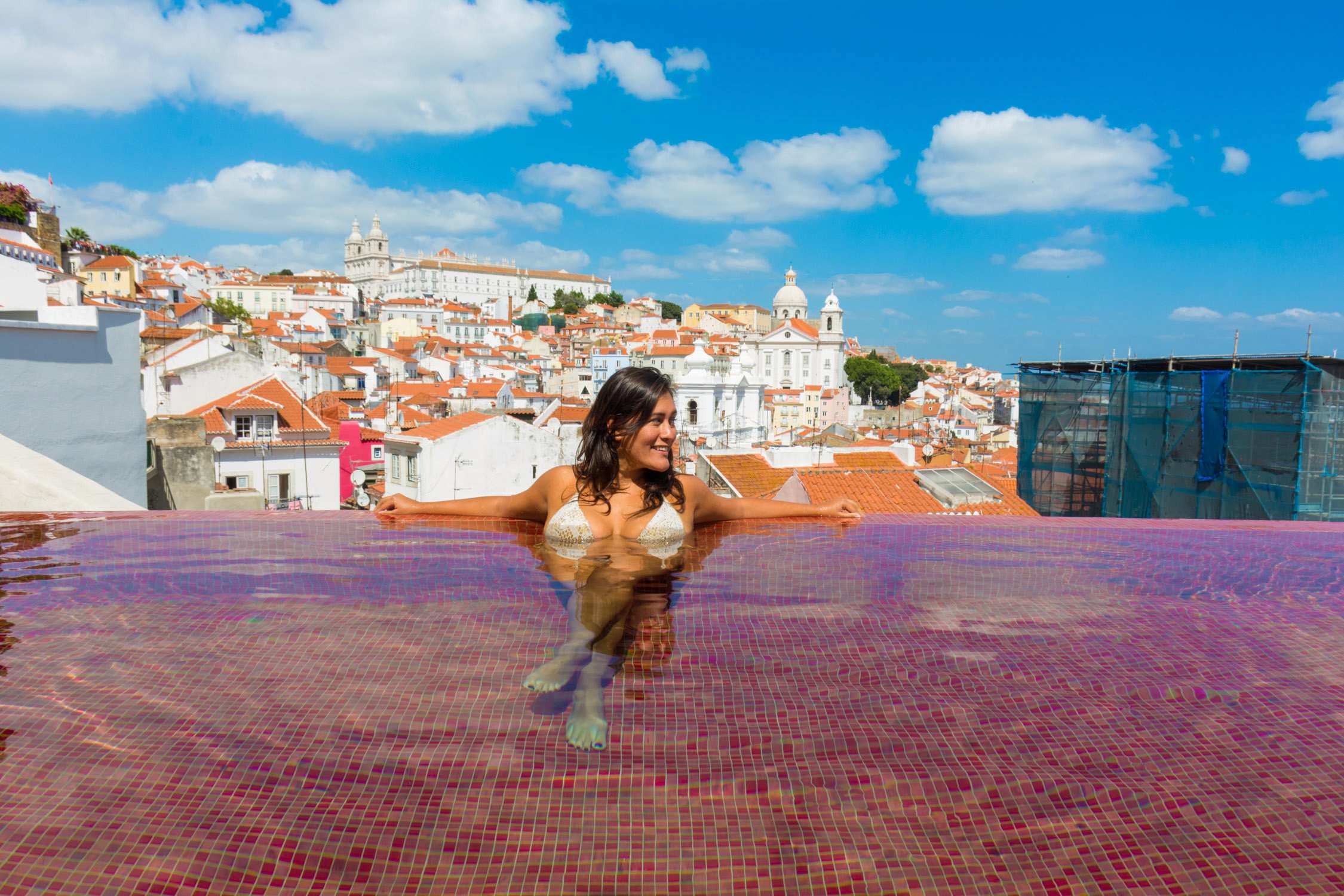 A woman in a bikini poses on a pink tiled terrace overlooking a coastal town with white buildings and blue roofs.
