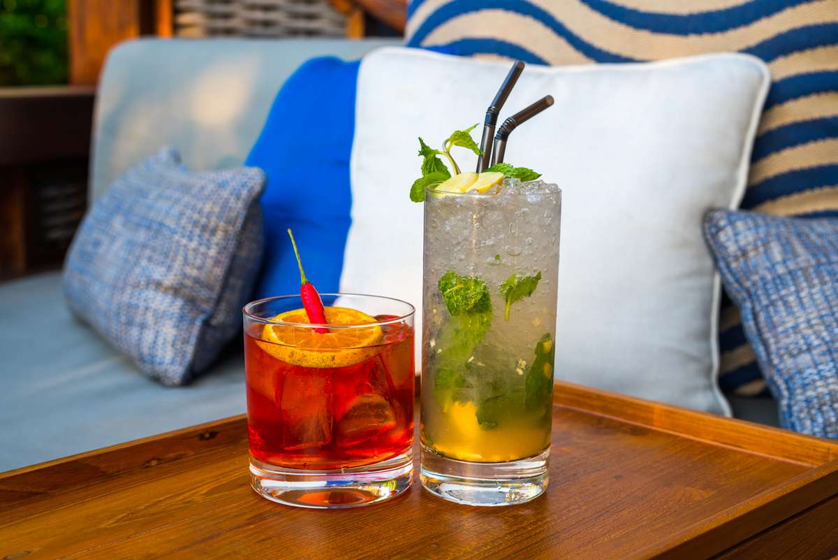 Two cocktails with mint and fruit garnishes sit on a wooden tray in front of blue and white striped cushions.