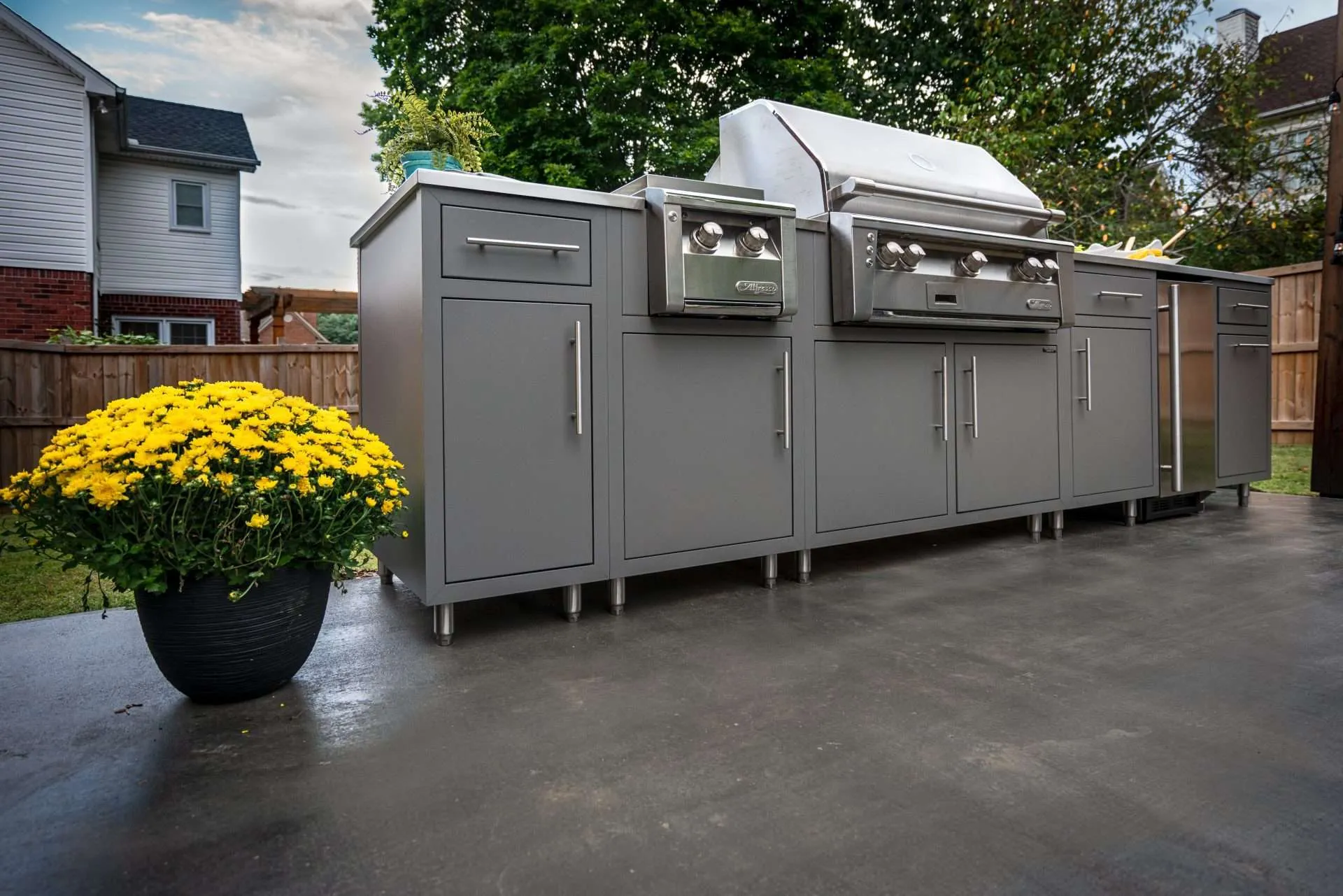 A modern outdoor kitchen featuring a gray cabinet system and a large stainless steel grill with side burners, set on a clean concrete patio. A large black planter with bright yellow flowers adds a pop of color, with a house and fenced yard in the background.