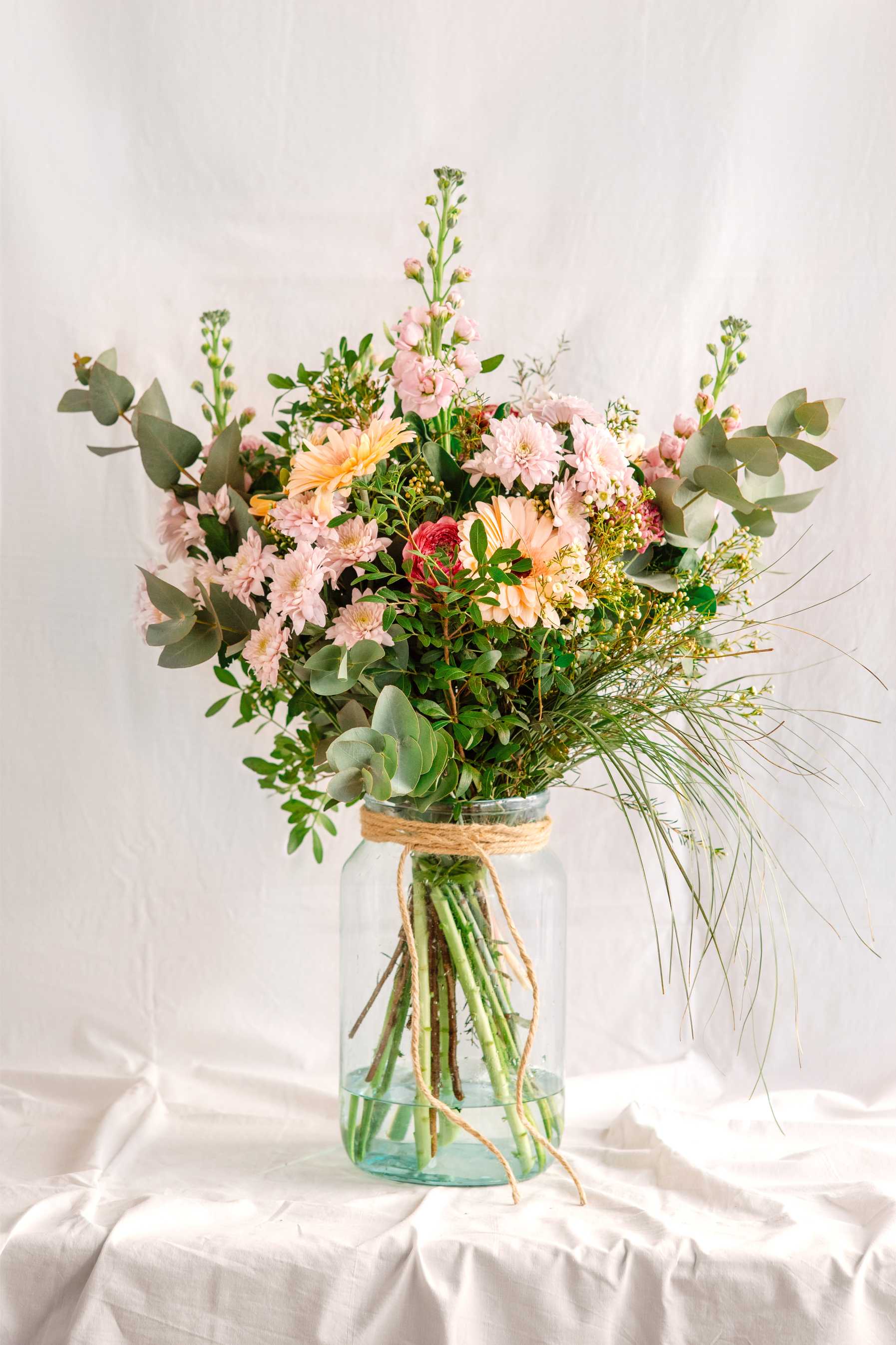 Ram de flors silvestres amb gerberes, crisantems, eucaliptus i flor de cera en tons suaus, presentat dins d’un gerro de vidre amb corda natural. Composició floral d’estil rústic i delicat.