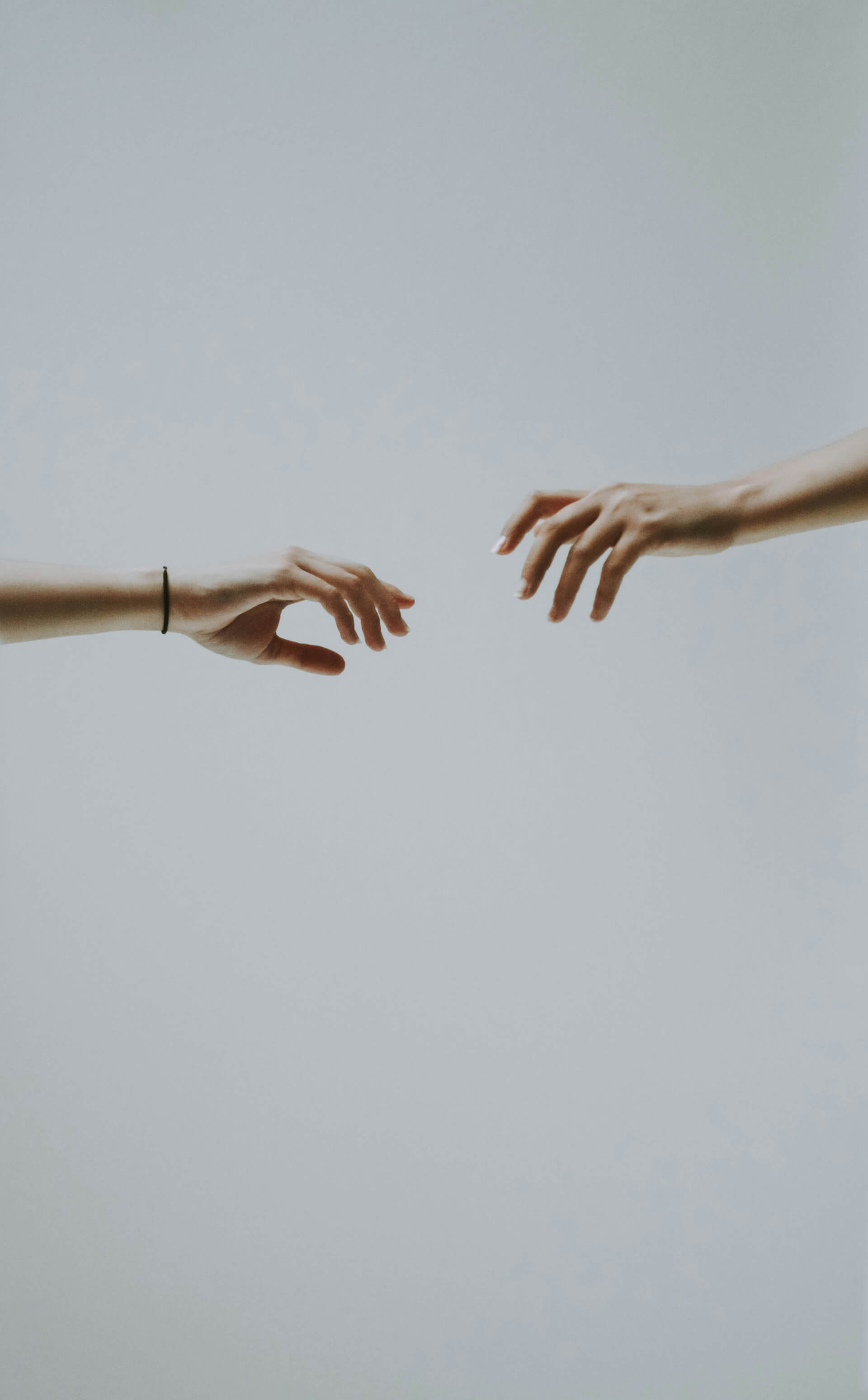 Two hands reach toward each other against a soft gray background, their fingers almost touching in a delicate and intimate gesture. One wrist wears a simple black hair tie, adding a subtle human detail to the minimalist composition.