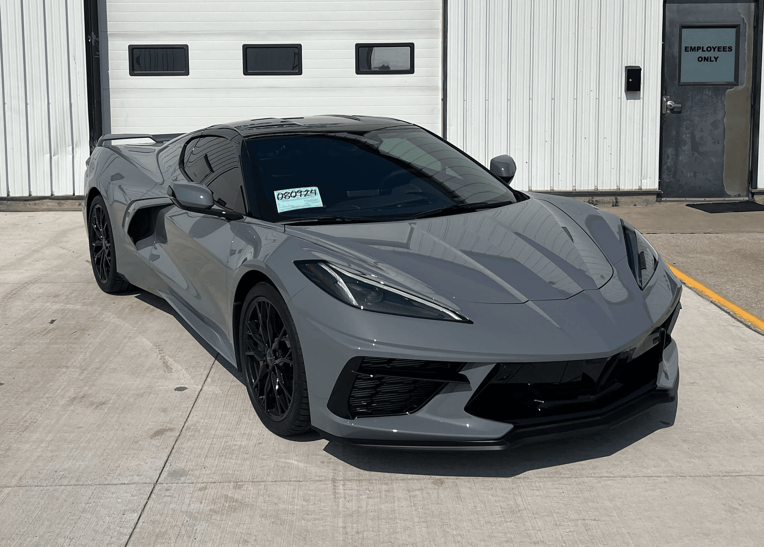 A sleek gray Chevrolet Corvette Stingray is parked on a concrete driveway in front of a white industrial building.