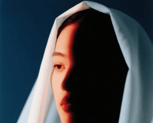 A person draped in a white cloth is partially illuminated by dramatic red lighting, creating a striking contrast against a dark blue background.