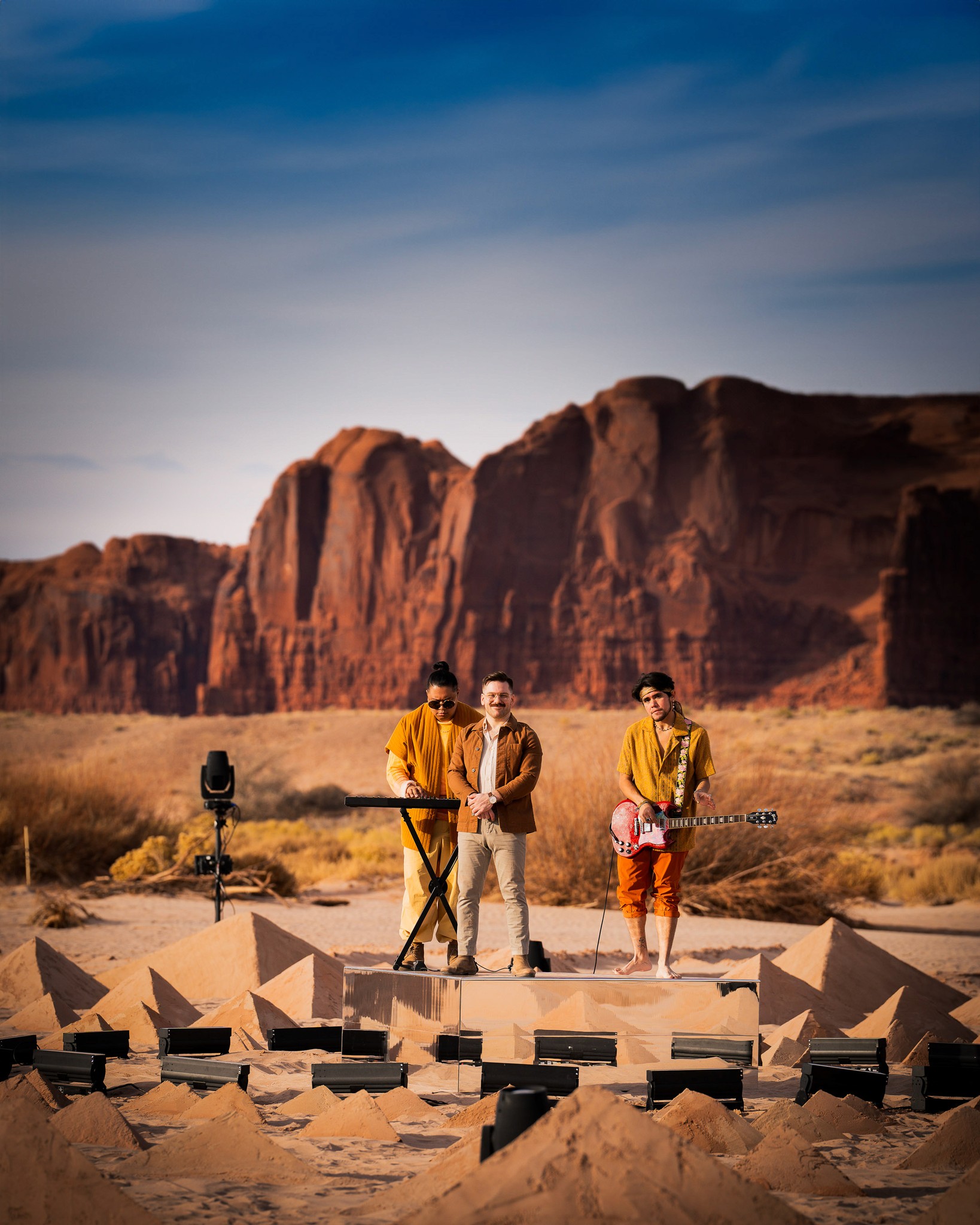 The band A R I Z O N A standing on a mirrored stage with pyramids made of sand in front of them and giant red rocks behind them