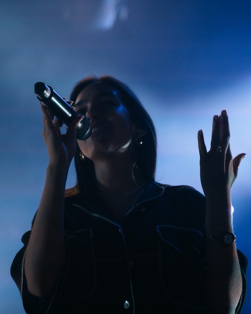 A monochrome image of performers on stage under dramatic lighting, with an audience visible in the foreground.