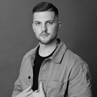 A man with short, tousled hair is wearing a dark blazer over a button-up shirt, standing against a neutral background, exuding a professional and confident demeanor.