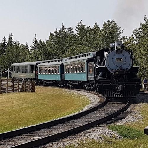 Um trem a vapor vintage com vagões verdes e azuis percorre uma trilha curva cercada por árvores e grama.