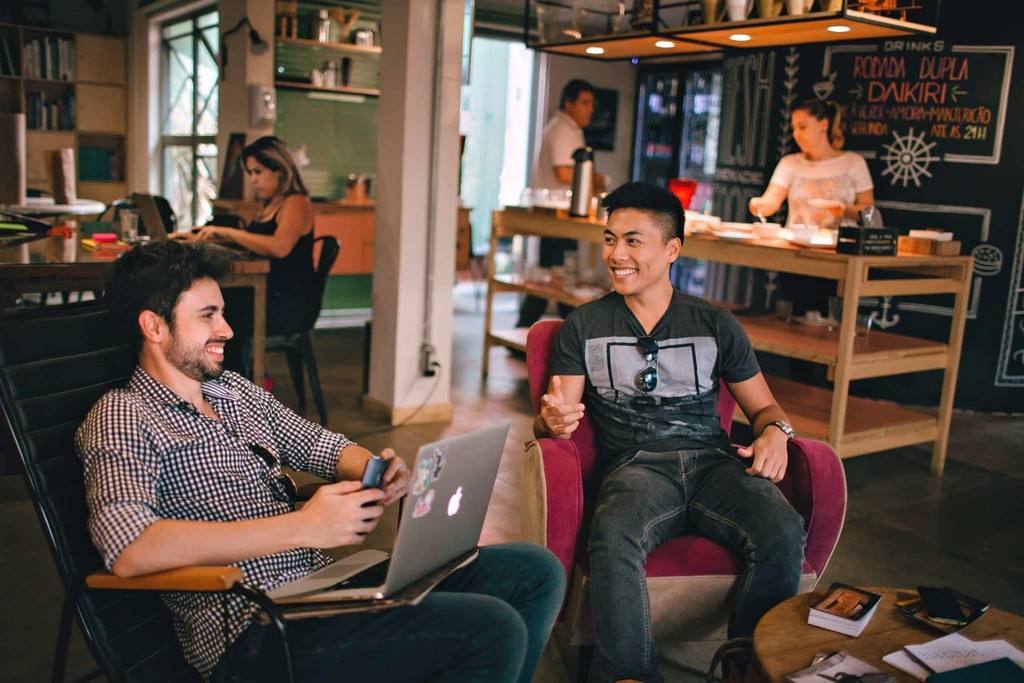 Smiling friends chatting in a modern café with laptops and coffee.