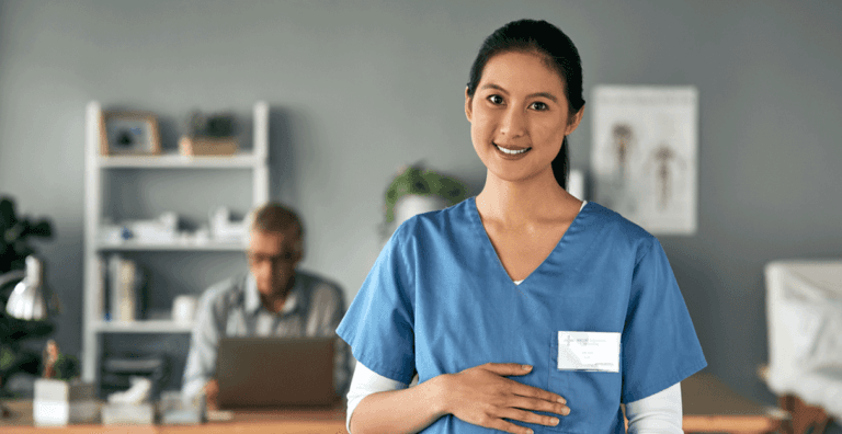 Smiling nurse in blue scrubs standing in a clinic with a doctor working in the background.