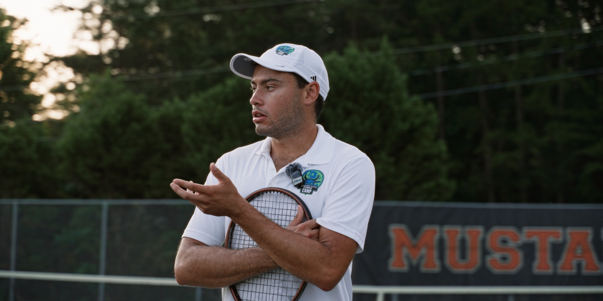 Coach de tennis en pleine discussion, raquette en main et logo du camp visible.