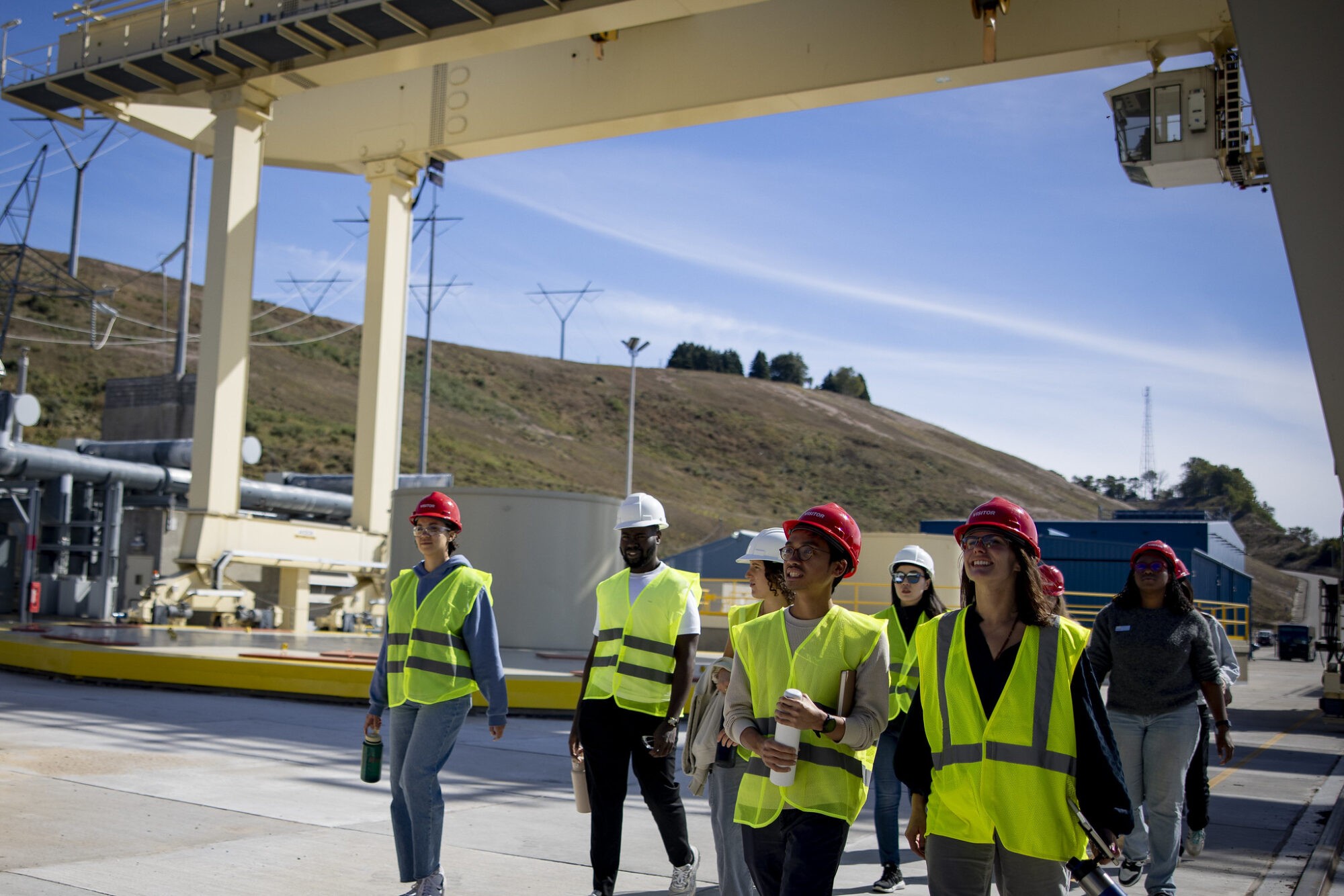 A group of workers in reflective vests discuss plans under a large construction structure on a sunny day.