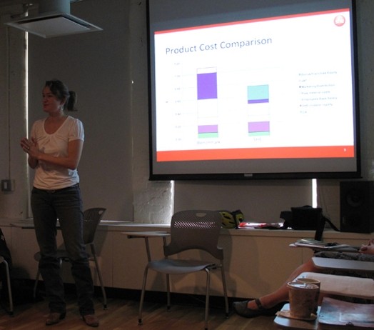 A photo of a woman giving while having behind her a projector screen that shows: product cost comparison.