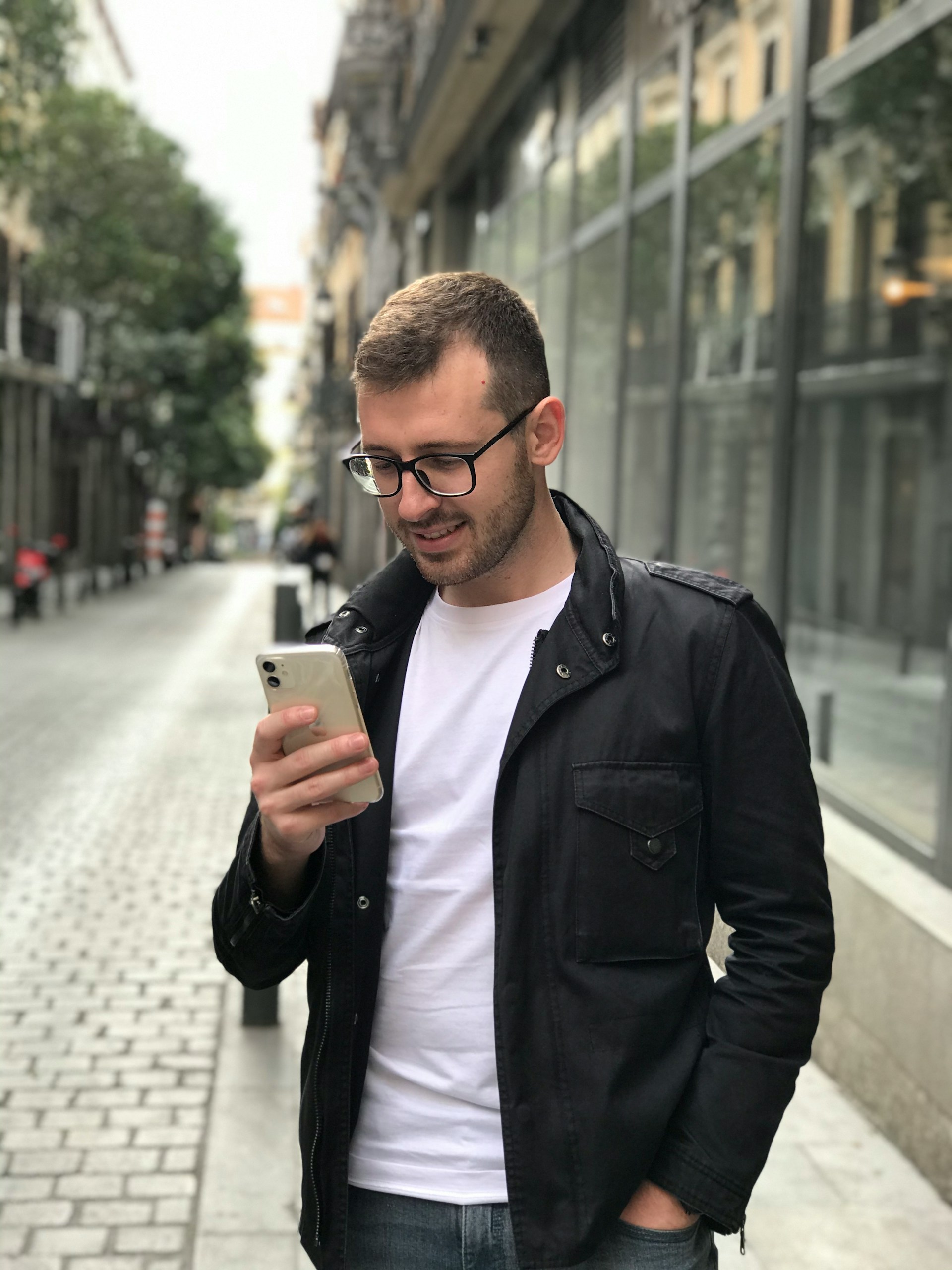 A man wearing glasses, a white t-shirt, and a black jacket is standing on a narrow city street, looking down at the smartphone he is holding.
