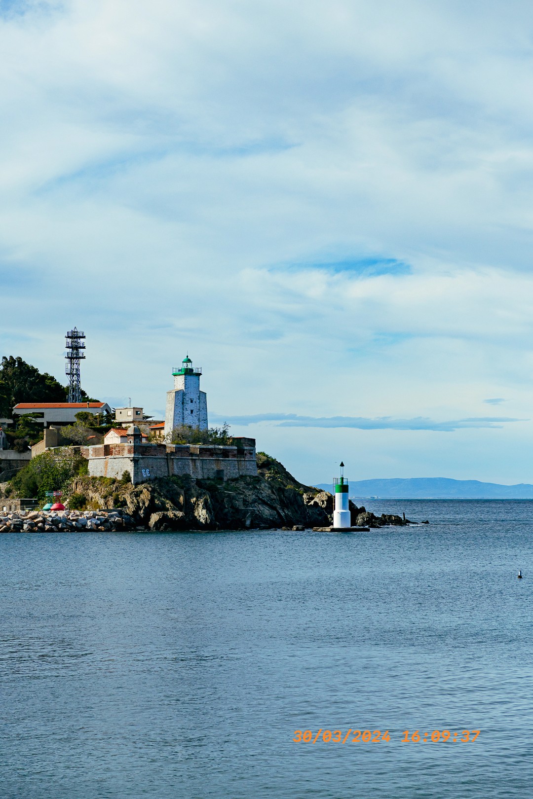 Phare blanc sur pointe rocheuse, petite balise verte en avant-plan.