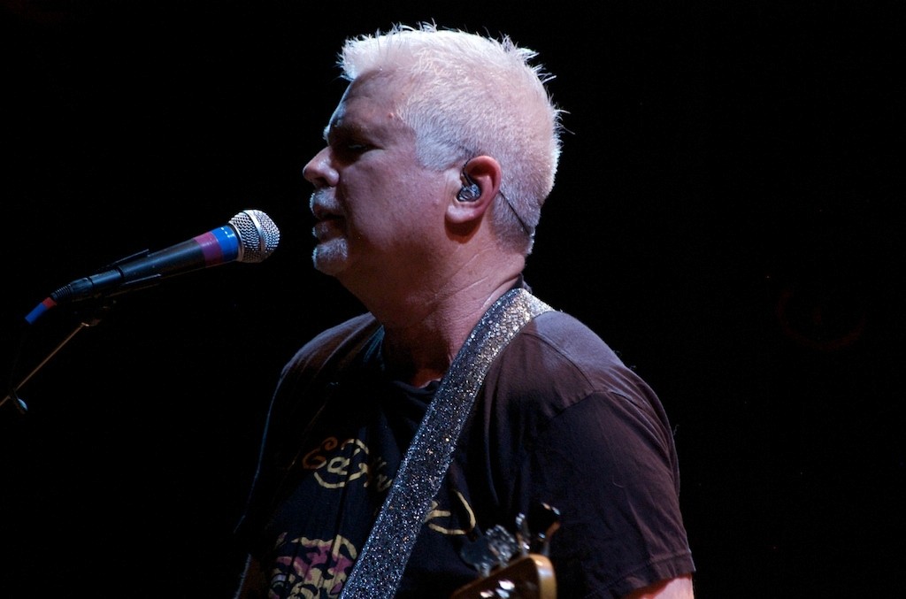 A musician sings into a microphone, illuminated against a dark background, with gray hair and a focused expression.