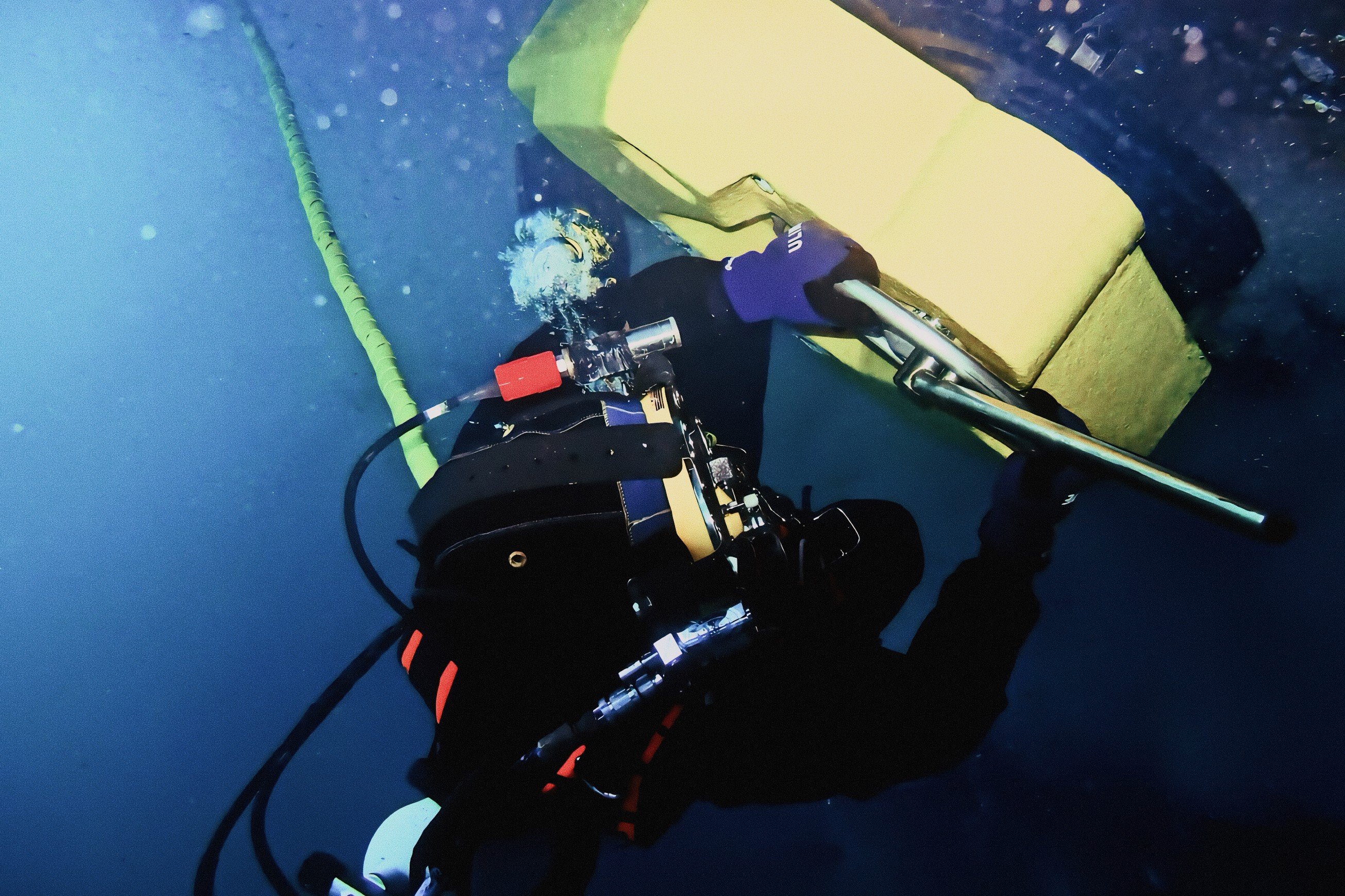 Commercial diver underwater using a hydraulic cleaning tool near a ship’s hull, working beside a large yellow fender with hoses visible in blue water.