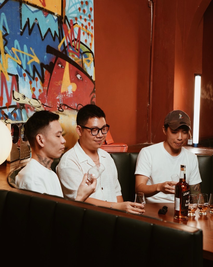 Three individuals seated at a bar with a bottle of Jameson Black Barrel whiskey, engaged in a tasting experience amid a colorful mural backdrop.
