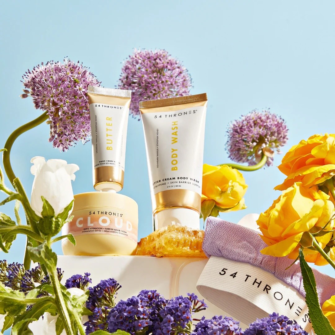 Hero composition of the 54 Thrones Cloud Cream, Body Butter, and Body Wash — styled among vibrant blooms to highlight the product’s natural, uplifting essence.