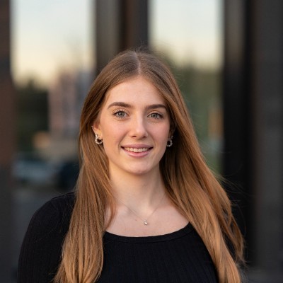 Eine junge Frau mit langen braunen Haaren lächelt im Freien, mit einem Gebäude und einem Sonnenuntergang im Hintergrund.