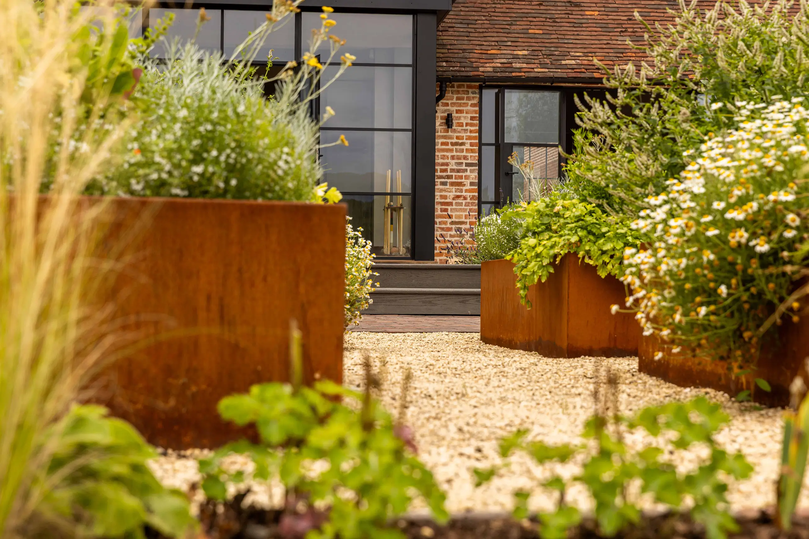 A garden scene featuring large planters with vibrant greenery, leading to a door in a brick wall.