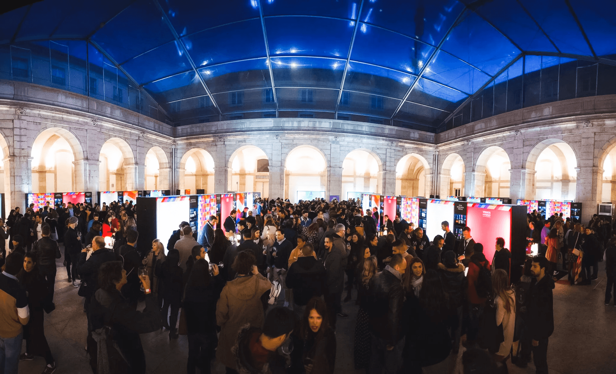 Crowded event beneath a blue-lit glass roof and stone arches at Pátio da Galé