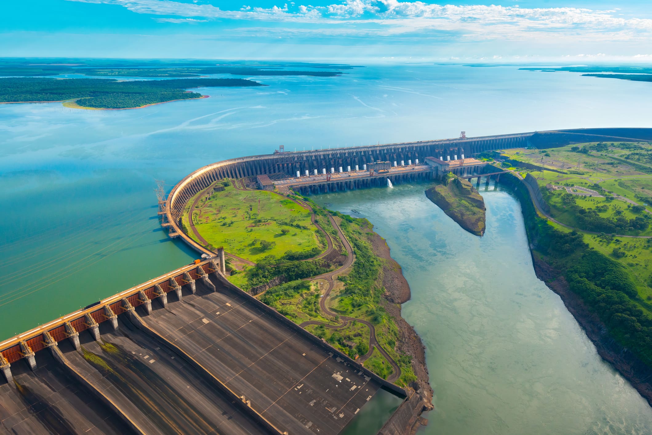 Aerial view of a vast hydroelectric dam with curved structure spanning a large river. Surrounding lush green landscape under a bright blue sky.