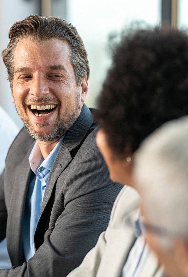 A man in a suit laughing warmly during a meeting, with colleagues visible in the foreground.