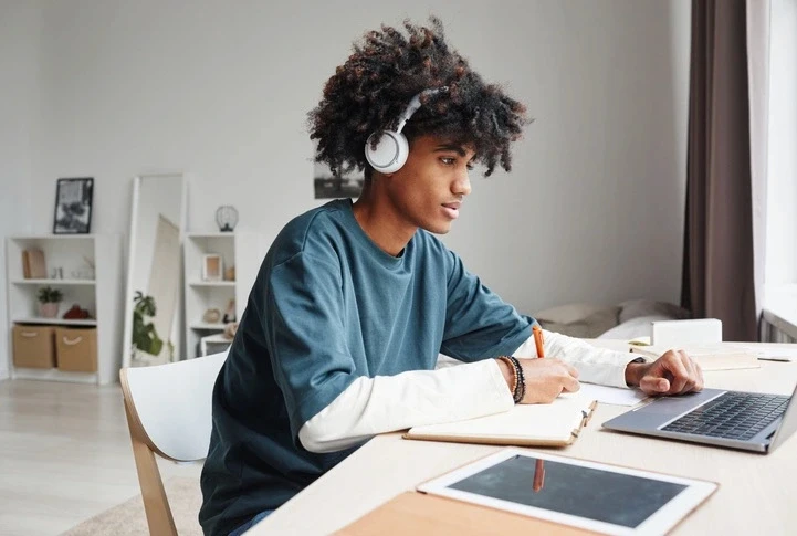 High school students in RISE Research or Ivy League summer programs studying STEM subjects; teen with headphones writing at desk with laptop and tablet.