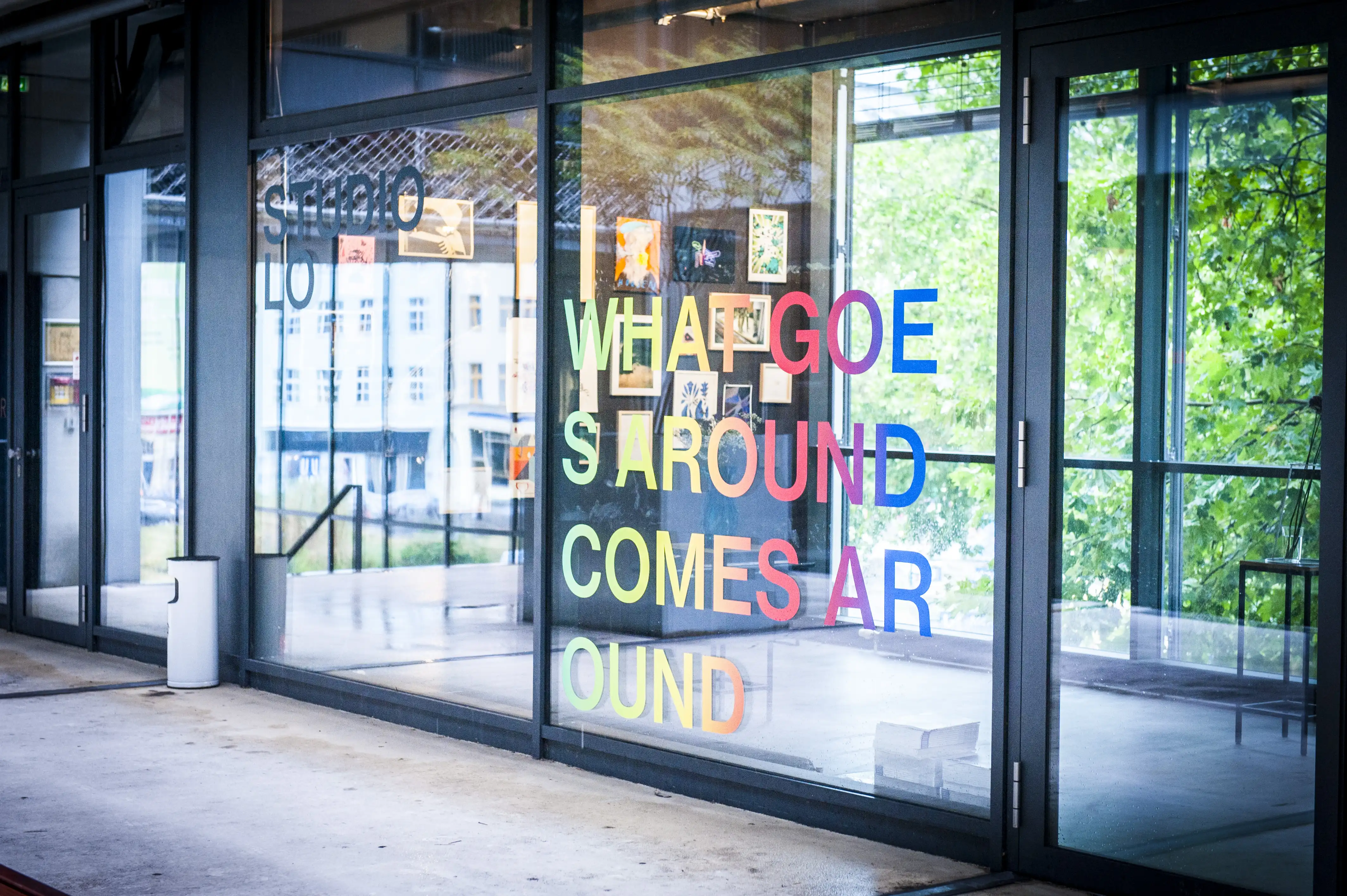 Glass entrance of the STUDIOLO gallery with multicolored What Goes Around Comes Around window lettering and framed artworks inside.