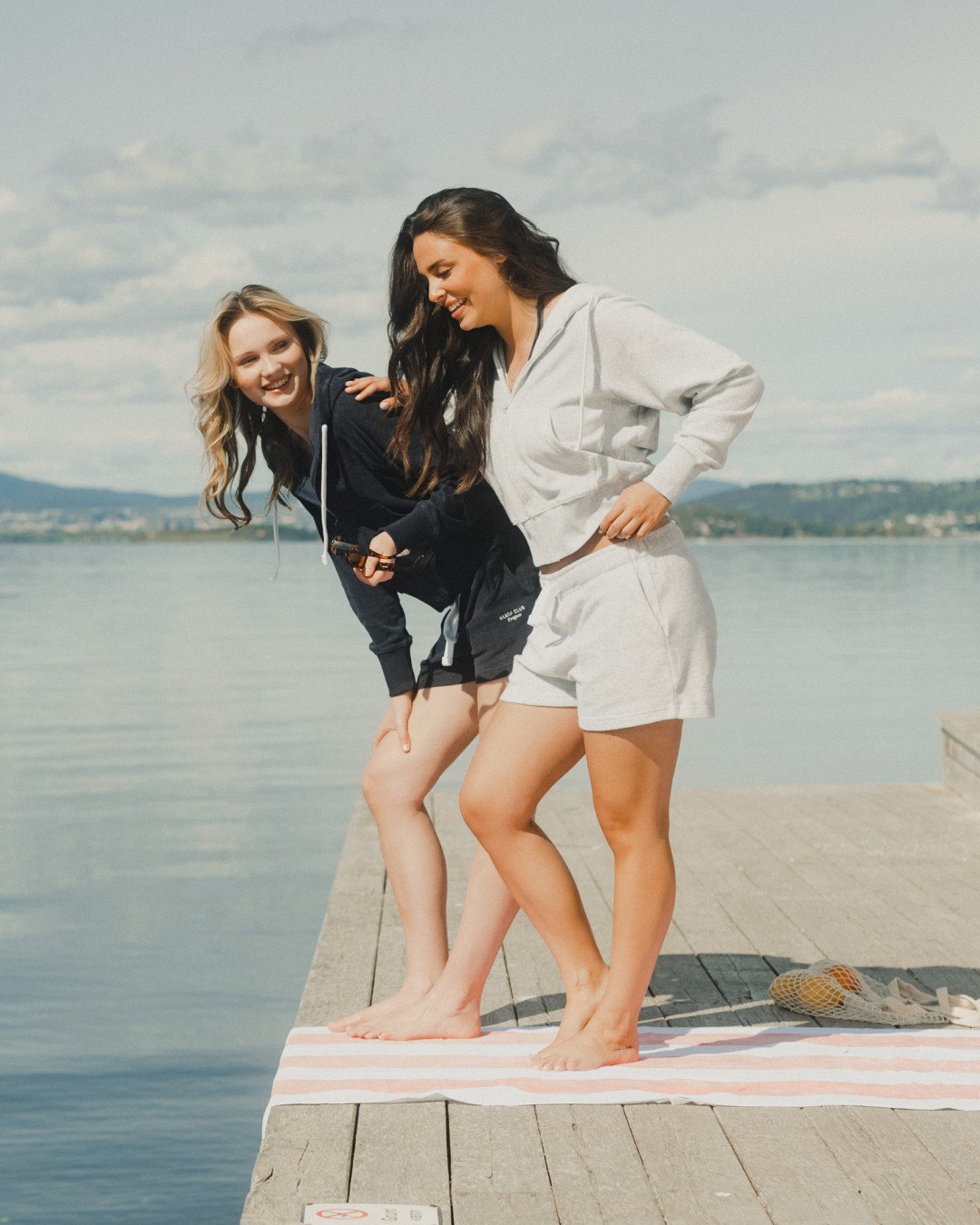 Two young women dressed in Russemerch ziphoods and shorts, standing on a dock by the water