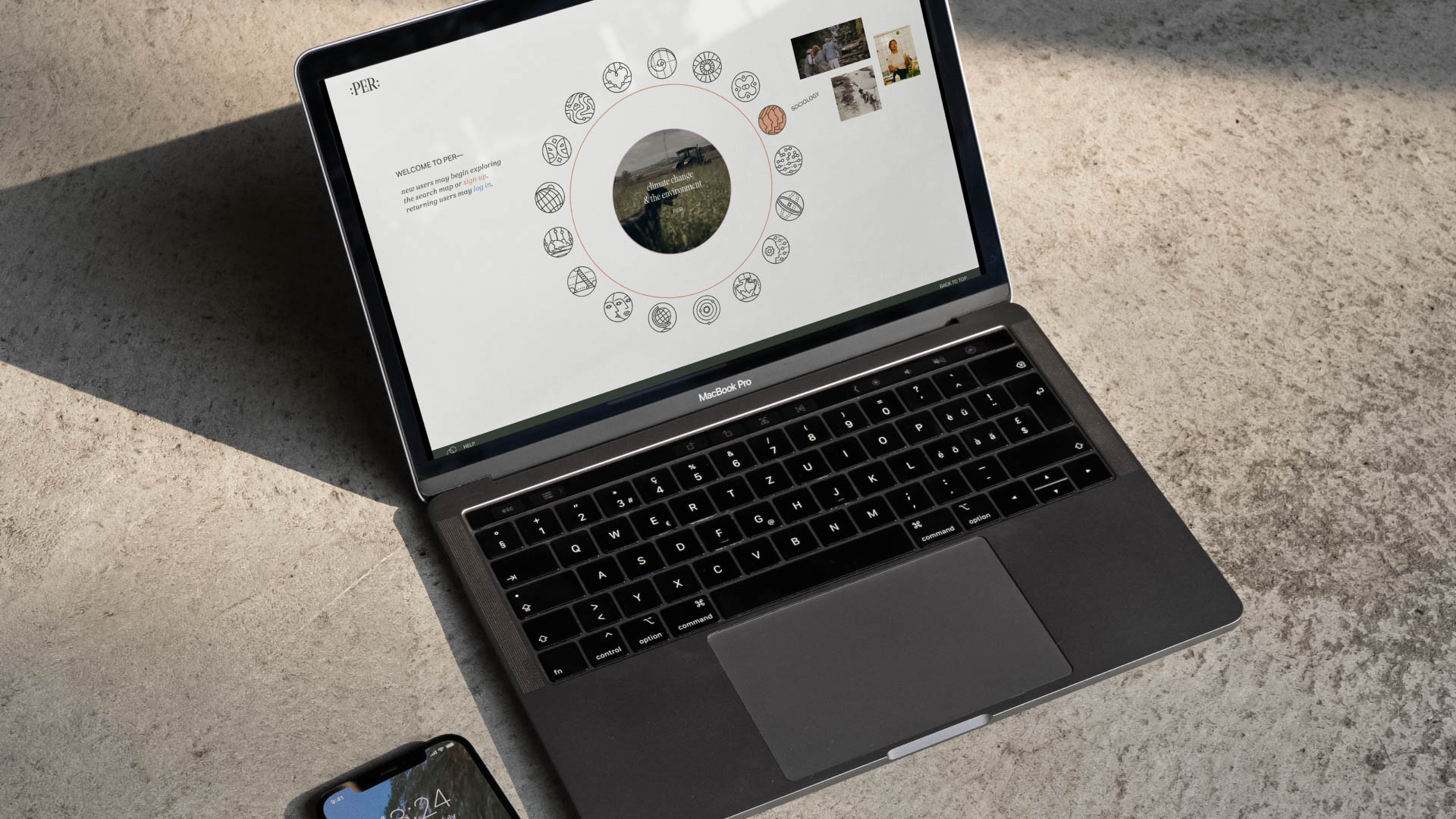 A black MacBook Pro laptop is open on a light gray speckled surface. The screen displays a circular design composed of small images and text, including the words “WELCOME TO FEB—” and “new year, new life, new beginnings.” A smartphone is partially visible in the lower left corner. The laptop’s keyboard and trackpad are dark gray or black.
