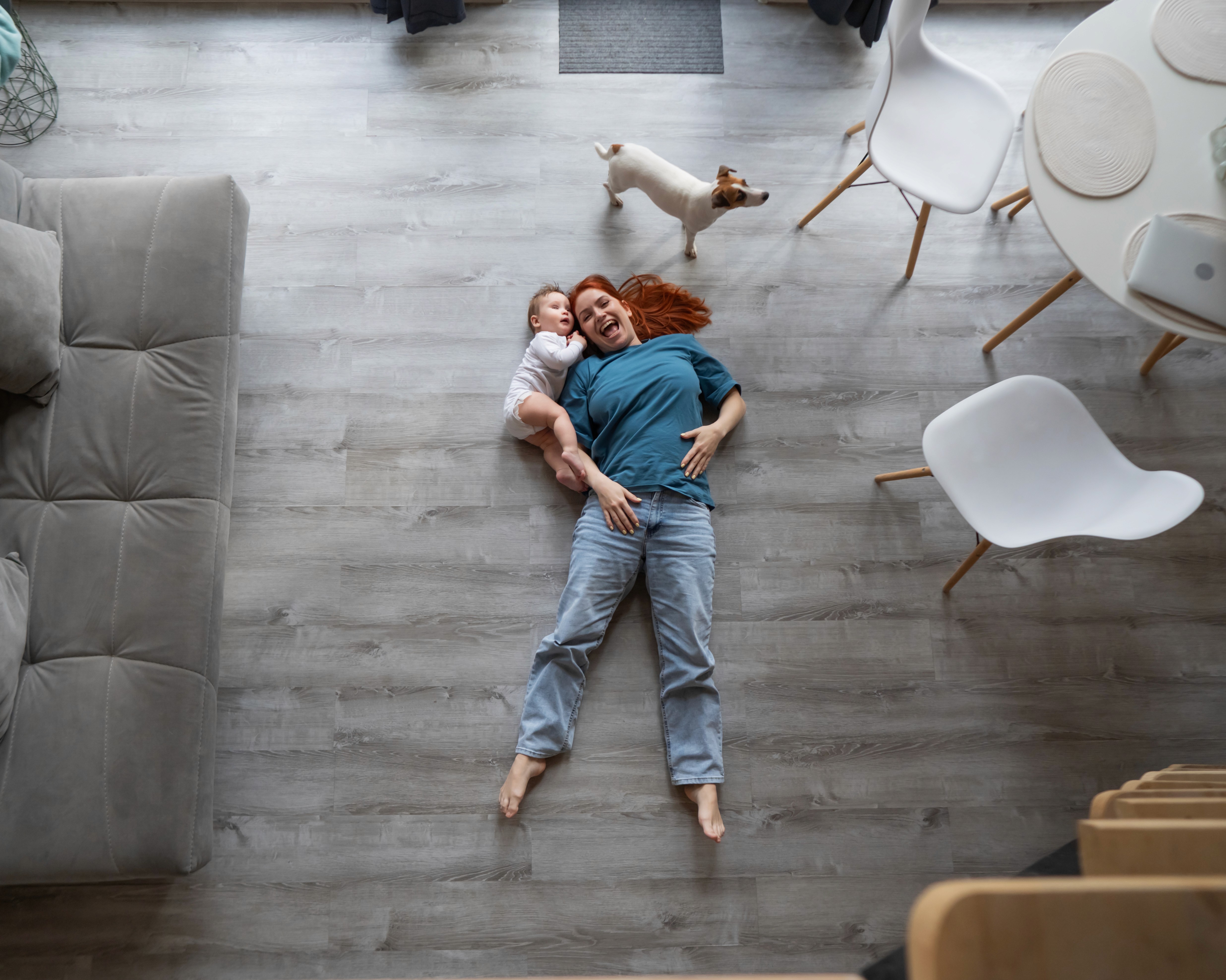 A mother relaxes on LUXO’s grey wood-look hybrid flooring with her baby and dog nearby, highlighting a safe, family-friendly, and easy-clean surface designed for modern Australian homes.