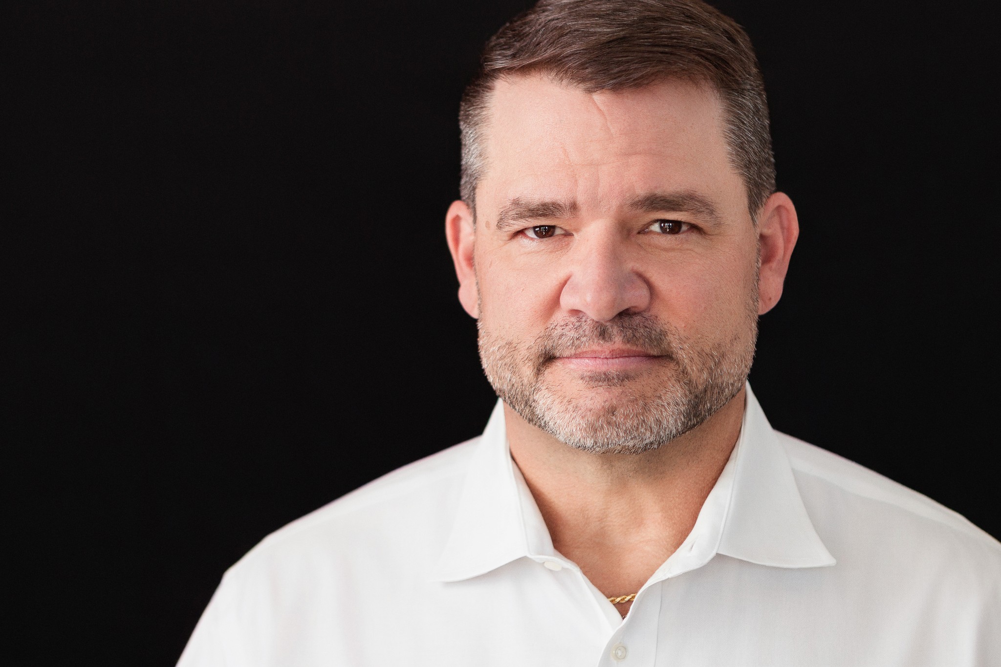 Executive branding portrait of a professional man in a white shirt against a black background, photographed with soft, directional studio lighting.