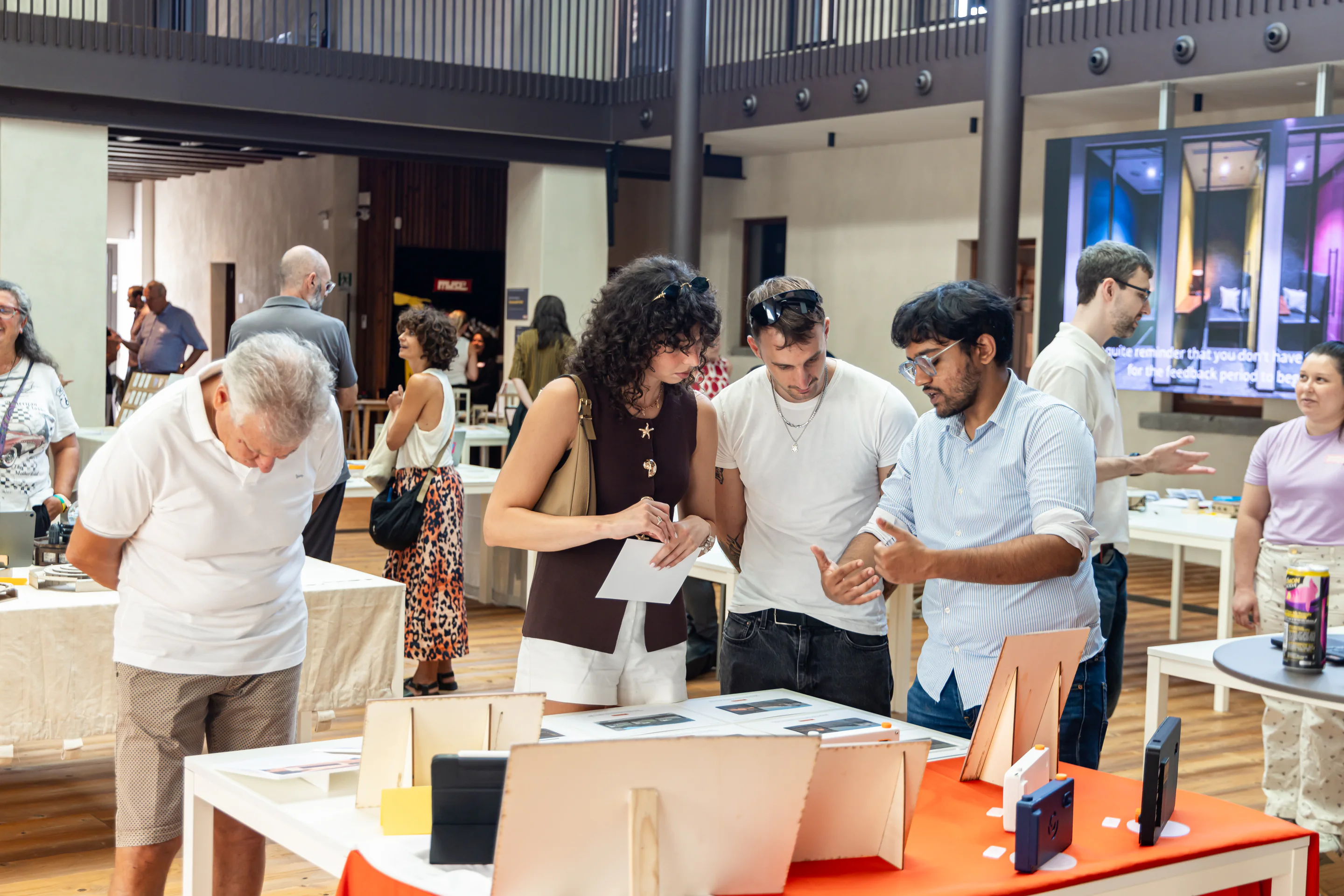 Three people discuss printed project boards at an exhibition table displaying camera prototypes.