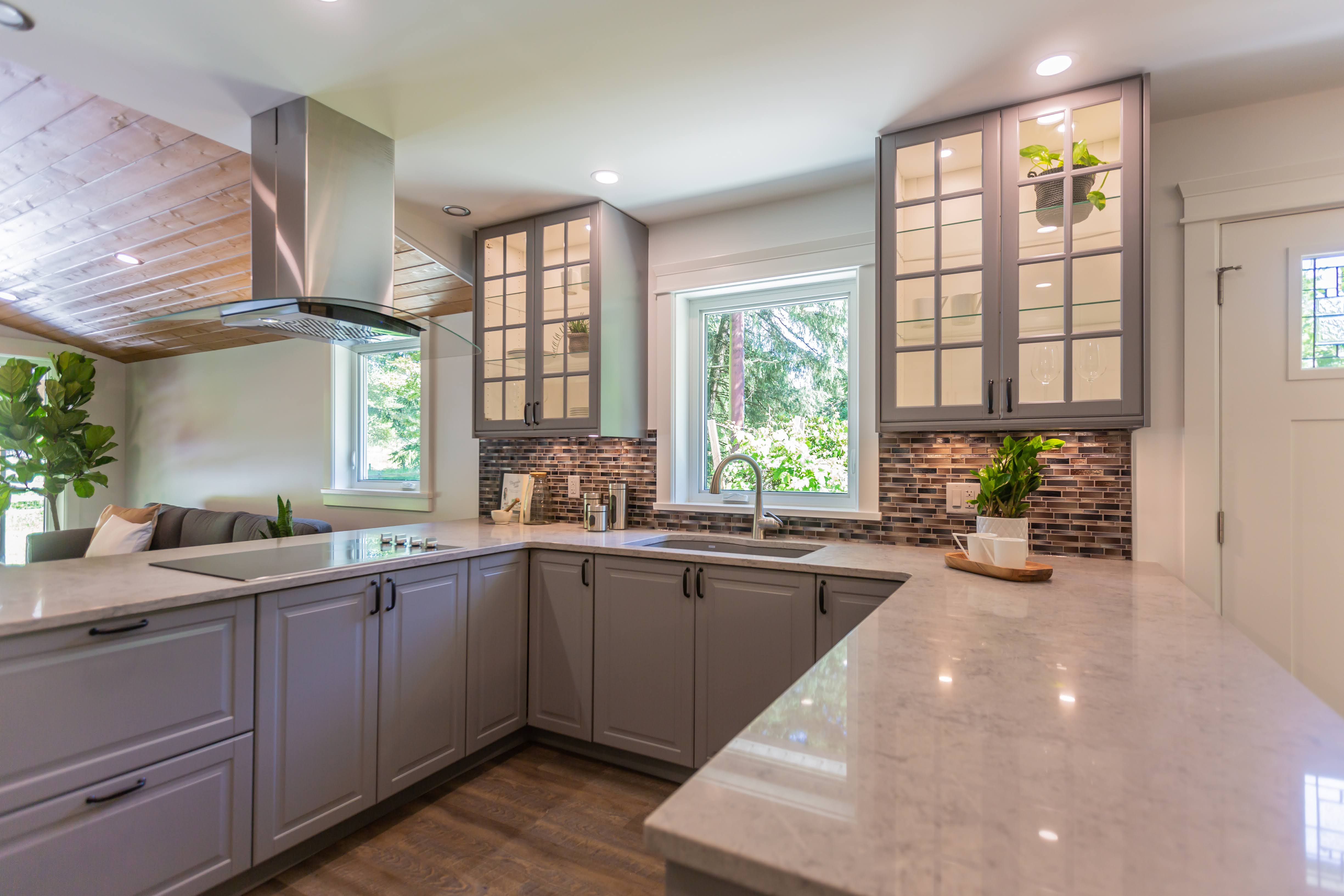 Contemporary U-shaped kitchen featuring light gray cabinetry, white quartz countertops, a stainless-steel range hood, and a mosaic tile backsplash beneath a window.