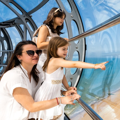 Three people in white attire observing a beach view from inside a glass observation pod; one girl points outside excitedly.