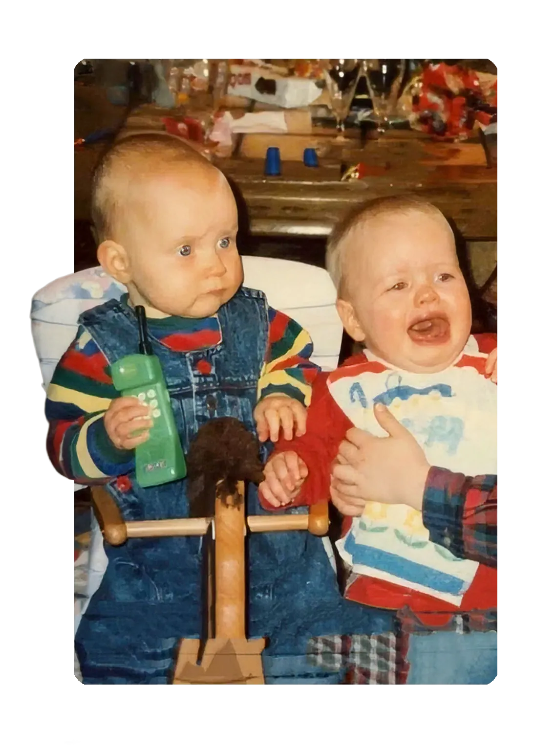 Zwei Babys im Hochstuhl, eines mit Spielzeugtelefon, eines weint. Kleinkinder Kindheit
