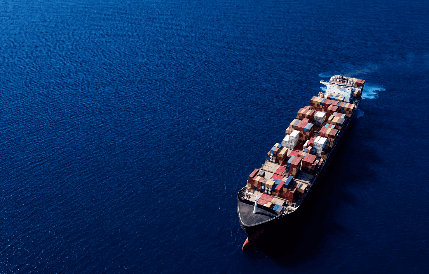 Aerial view of a large cargo ship at sea, loaded with colorful containers, sailing through deep blue waters, evoking a sense of global trade and vastness.
