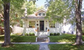 A charming, single-story Craftsman-style house with white siding and a small front porch, framed by two large trees with green foliage on either side. A concrete walkway leads from the sidewalk up a few steps to the front door, passing through a well-manicured lawn under a sunny sky.