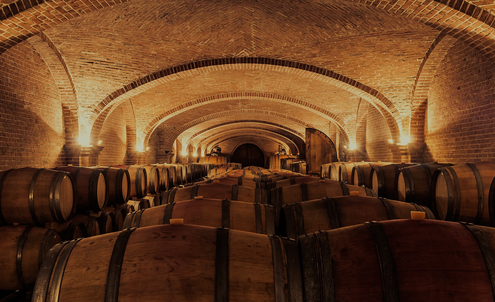 Brick wine cellar filled with oak barrels under arched ceilings, warmly lit by wall sconces.