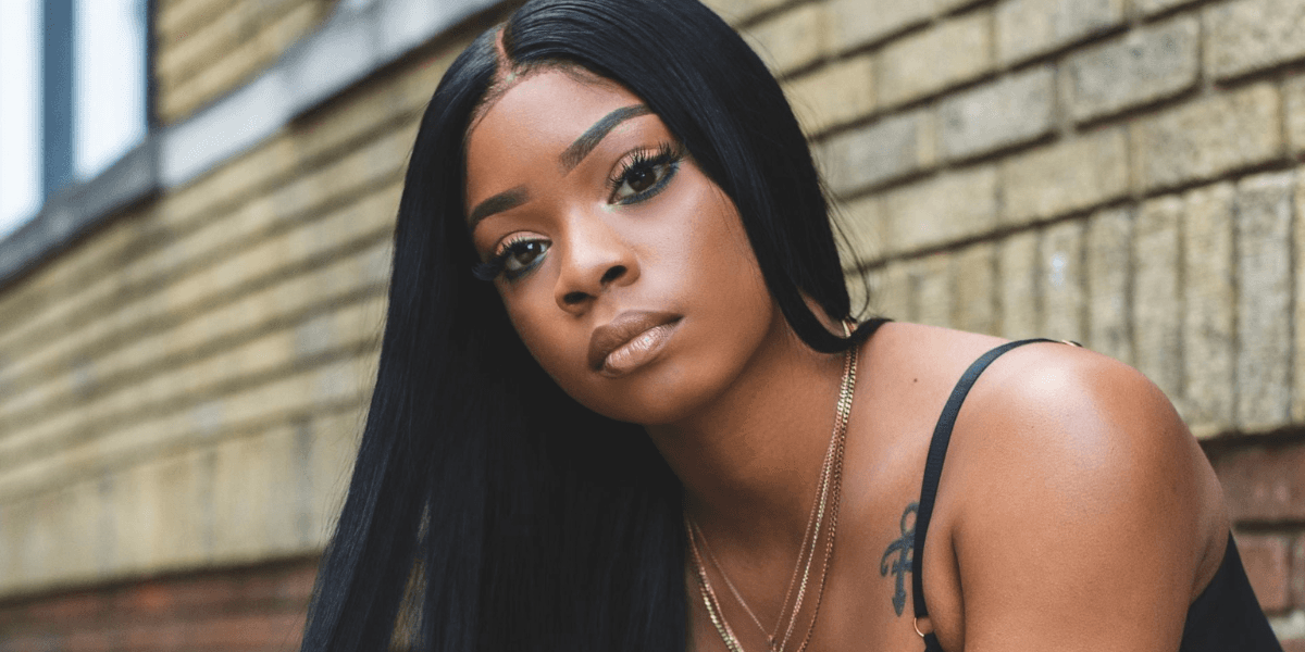 Young woman with long black hair and a confident expression poses against a brick wall. She wears a black top and gold necklaces, conveying strength and style.
